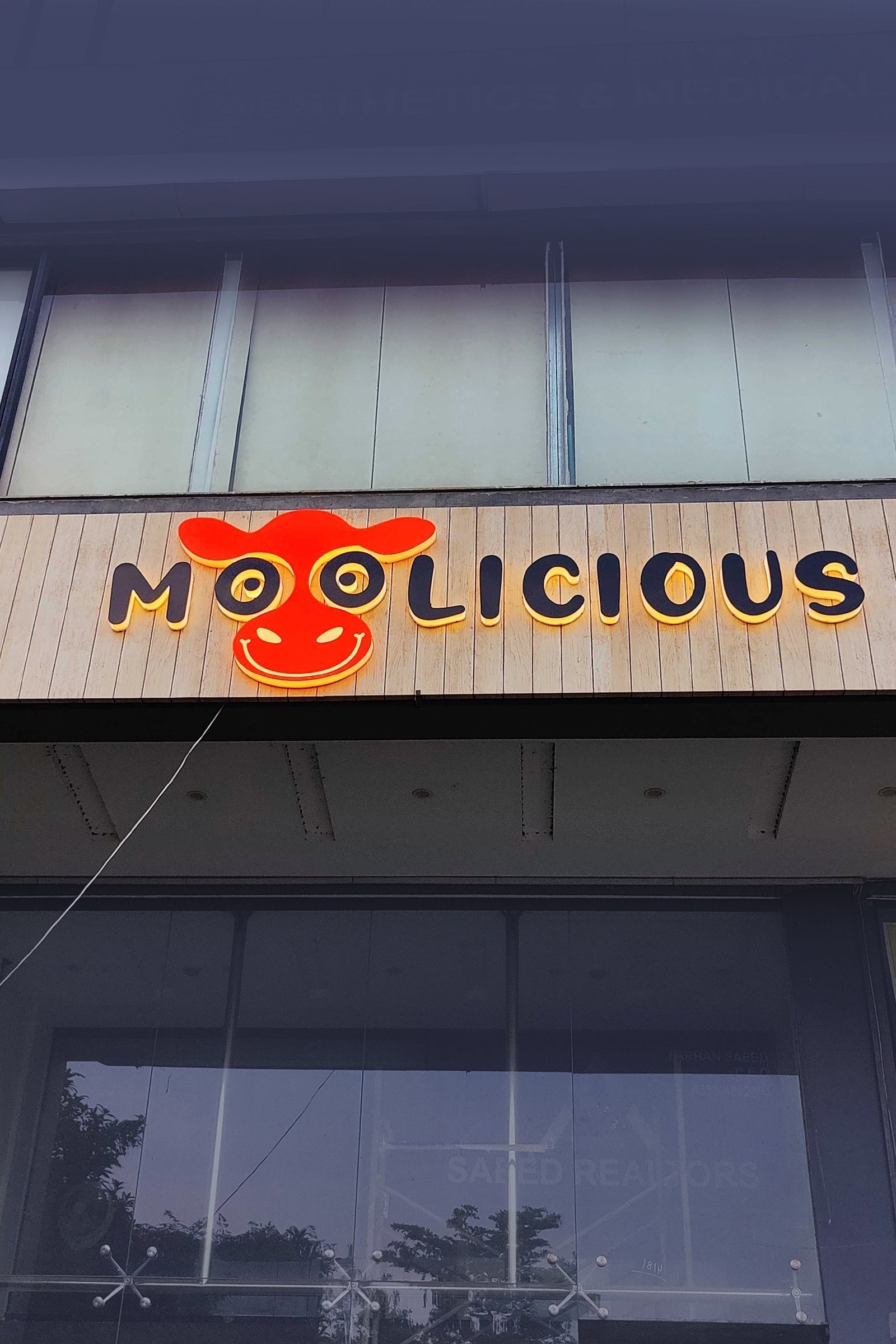 Illuminated business sign for Moolicious with a red cow face logo on storefront facade