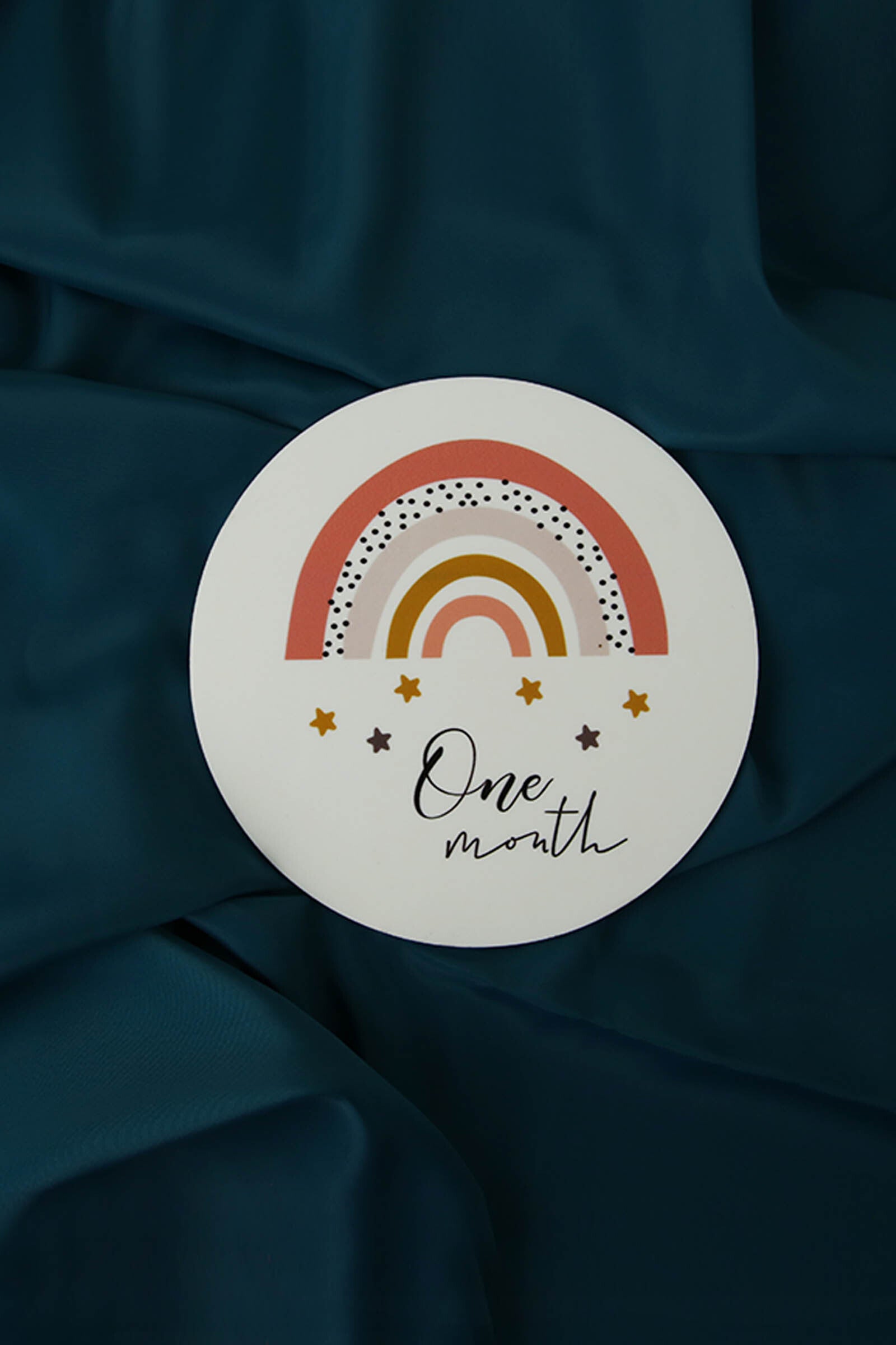 Round milestone plaque with rainbow and stars design reading 'One month' on blue fabric
