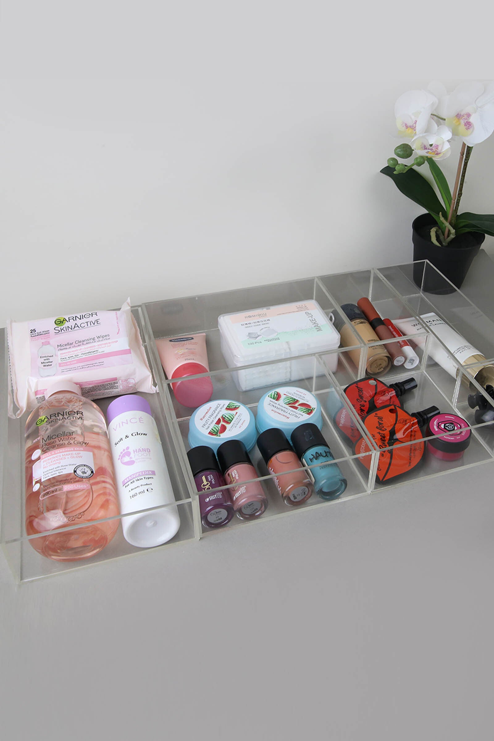 Clear acrylic makeup organizer holding cosmetics and skincare, with a potted orchid nearby.